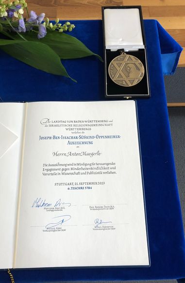 Auf einem Tisch mit blauem Tischtuch liegt die Verleihungsurkunde zur Oppenheimer-Auszeichnung für Anton Maegerle sowie rechts darüber in einer offenen Schachtel die goldene Oppenheimer-Medaille.