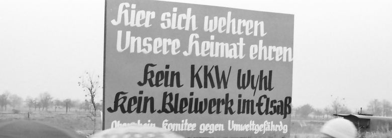 Schwarz-weiß-Fotografie einer Kundgebung gegen den Bau eines Atomkraftwerks im badischen Wyhl, 10. November 1974. Oben mittig ein Schild mit der Aufschrift: Hier sich wehren. Unsere Heimat ehren. Kein KKW Wyhl. Kein Bleiwerk im Elsaß. [Oberrheinisches] Komitee gegen Umweltgefährd[un]g. Im Vordergrund unten fünf vorbeilaufende Personen, zwei davon mit Regenschirm.